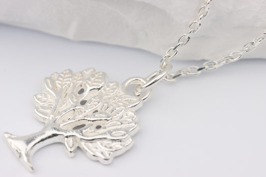 May include: A silver tree pendant necklace. The pendant features a detailed, stylised tree with a curved trunk and leafy branches. The necklace chain is delicate silver, with a simple clasp. The background is a soft white.